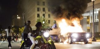 Two Black protesters on a motorbike, one of them raises a fist, in front of a burning car in a dark city street.