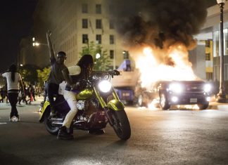 Two Black protesters on a motorbike, one of them raises a fist, in front of a burning car in a dark city street.