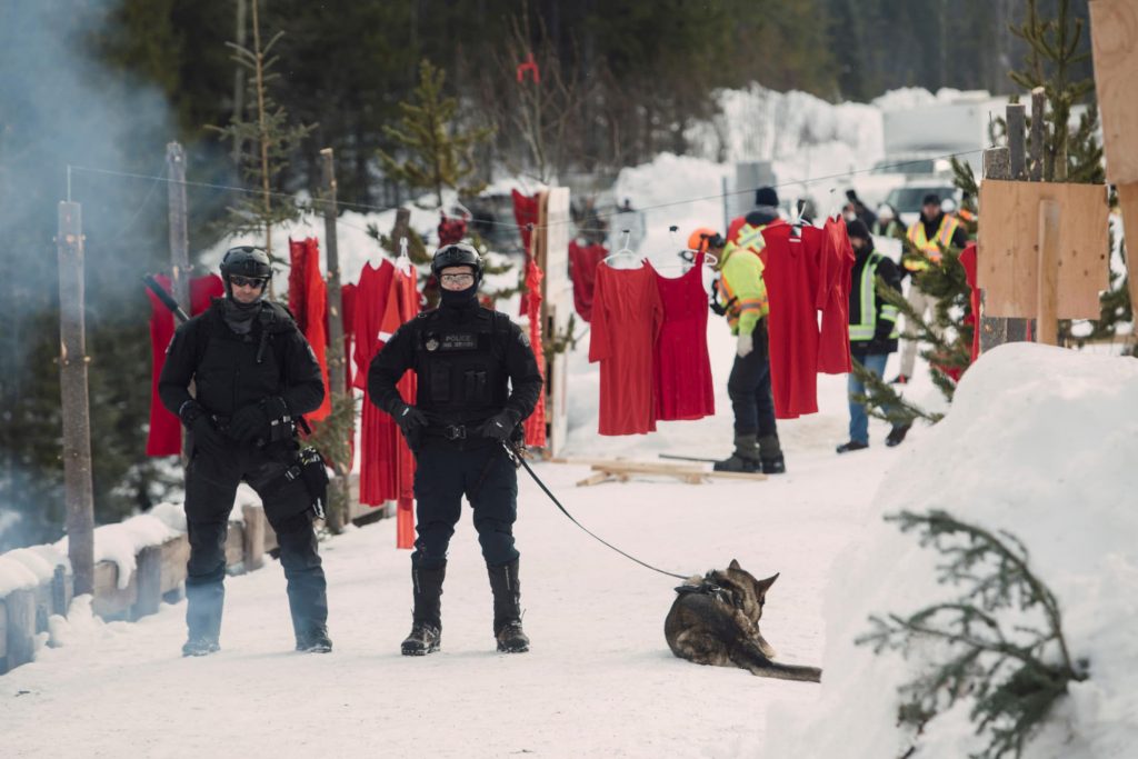 RCMP officers patrol the Unist'ot'en camp with dogs after a raid. In the background, red dresses hang, symbolising Indigenous women missing and murdered under colonial occupation.