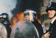 Three police officers in riot gear stand infront of a burning building on 31st of March 1990