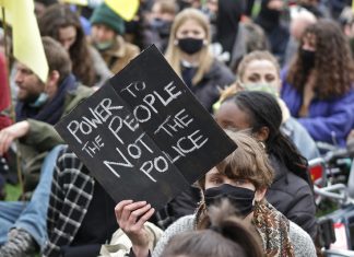 Image shows a woman holding up a placard reading Power to the People Not the Police with a backdrop of other women in the crowd.