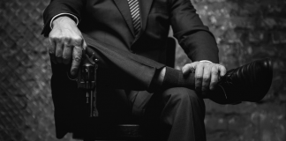 A shadowy figure in a suit holds a gun while sitting on a chair