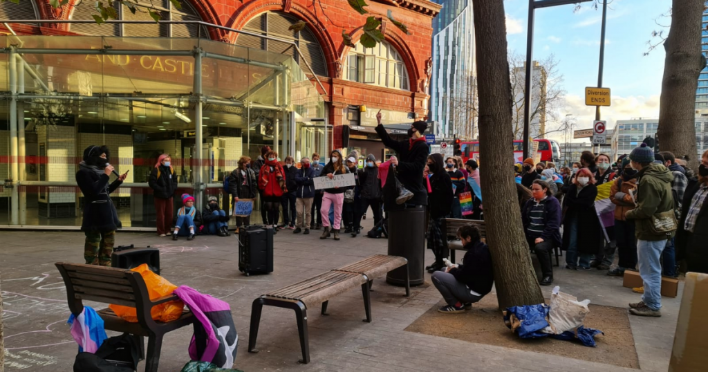 На фото изображены протестующие, собравшиеся вокруг выступающего у выступающего возле NHS England и станции метро Elephant and Castle на демонстрации 4 декабря 2021 года, организованной блоком действий трансгендеров.