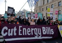 Image shows around 10-15 people at the front of a crowd raising their fists and holding up a purple cloth banner reading 'Cut Energy Bills'