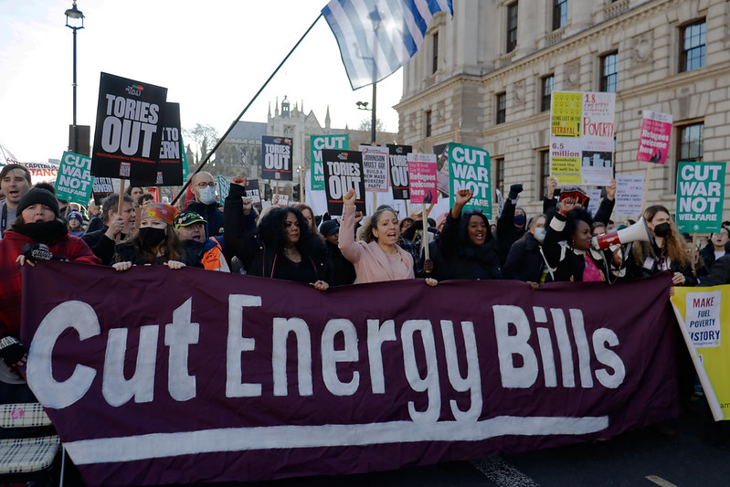 На изображении видно, как около 10-15 человек впереди толпы поднимают кулаки и держат фиолетовый тканевый плакат с надписью «Сокращение счетов за электроэнергию».