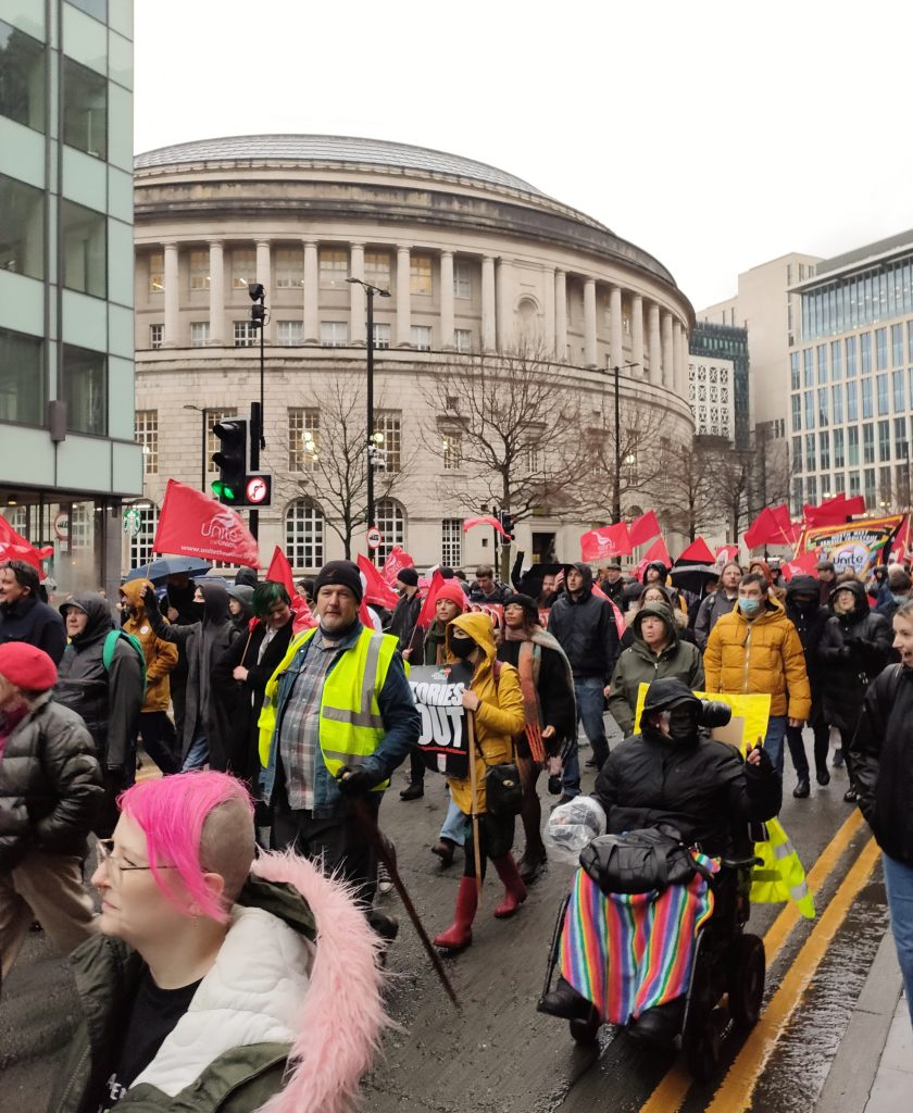 L'image montre environ 50 des 500 manifestants lors de la manifestation du coût de la vie de Manchester en passant devant l'emblématique bâtiment rond de la bibliothèque centrale.  Il y a de nombreux drapeaux Unite, des drapeaux rouges et une personne tient une pancarte «Tories Out».