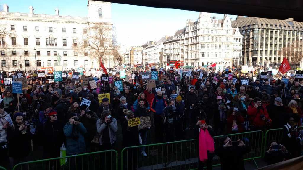 The image is the view from the stage at the London cost of living demonstration - a sea of placards, faces and a sunny sky in the background.