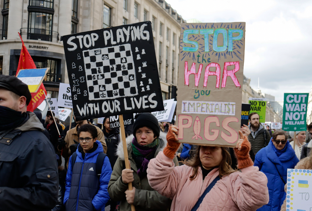 Crowd with placards. One has chess board and 'stop playing with our lives'. Another reads 'Stop the war your imperialist pigs'