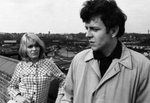 Screenshot is in black and white, showing a man in the foregound wearing a light-coloured coat, looking wistfully to his right, while behind him, a blonde woman stands looking up at him, unsmiling. She leans against a wall, which disappears to the left of the photo, against a backdrop of English scenery.