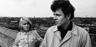 Screenshot is in black and white, showing a man in the foregound wearing a light-coloured coat, looking wistfully to his right, while behind him, a blonde woman stands looking up at him, unsmiling. She leans against a wall, which disappears to the left of the photo, against a backdrop of English scenery.