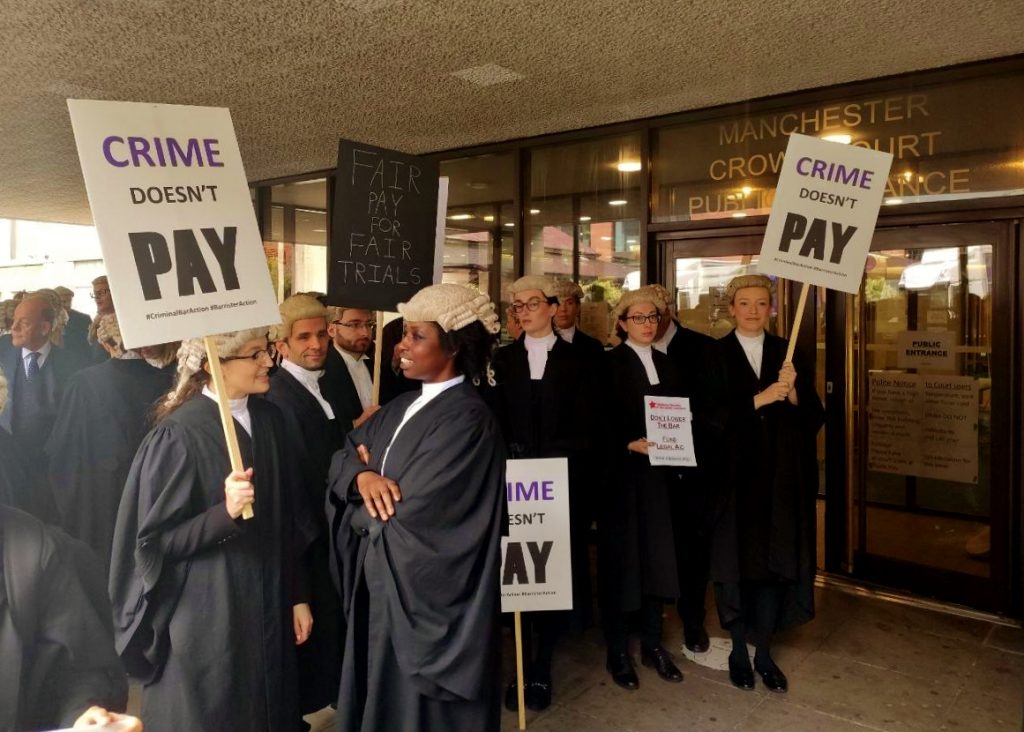 Photo taken outside Manchester Crown Court shows barristers in full robes on strike, holding placards reading 'Crime Doesn't Pay', 'Fair Pay for Fair Trials' and 'Legal Aid: Don't Lower the Bar'.