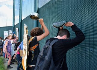 Image shows a high green fence lined with protesters banging metal items against it.