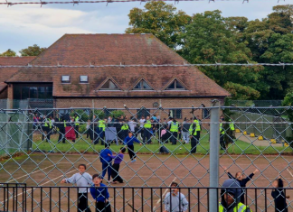 A photo of Manston camp from 30 October 2022, showing a large number of people fenced in with barbed wire and many security guards, with children running towards the barbed wire fence in front of the camera.