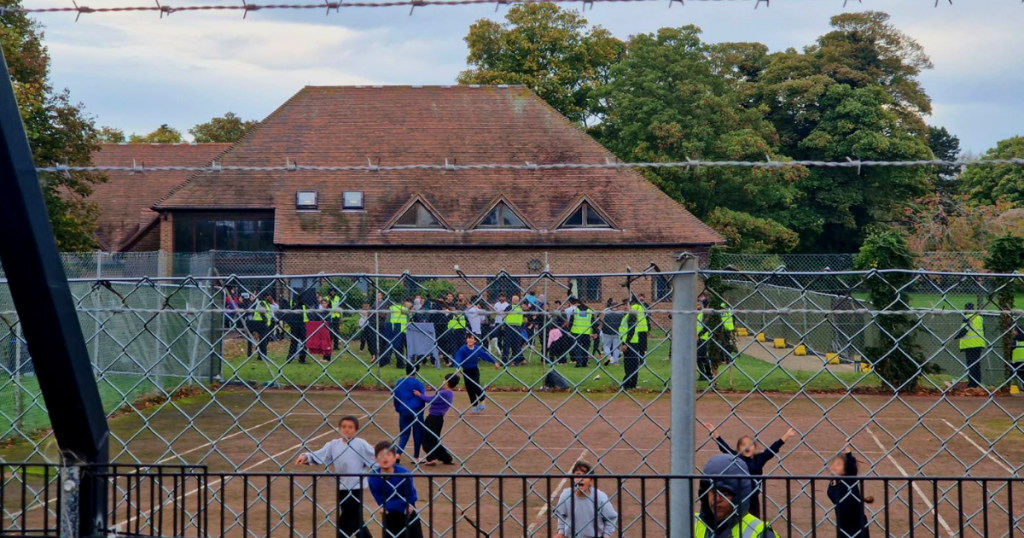 Фотография лагеря Мэнстон от 30 октября 2022 года, на которой видно большое количество людей, огороженных колючей проволокой, и множество охранников, а дети бегут к забору из колючей проволоки перед камерой.