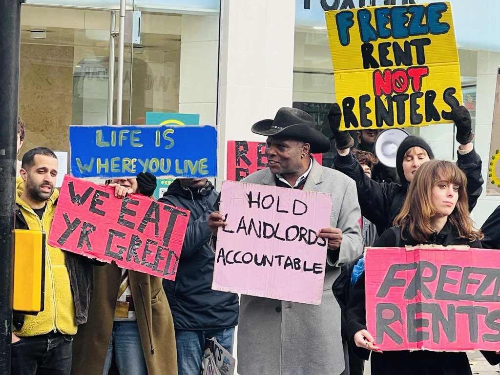 На изображении показана группа протестующих с плакатами «Жизнь там, где ты живешь», «Привлечь к ответственности владельцев домов» и «Заморозить арендную плату, а не арендаторов».