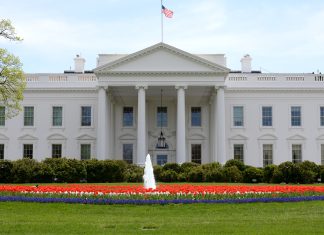a picture of the White House, with a United States flag flying on top, and red and blue flowers and a fountain below.
