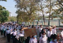 Teachers in Myanmar march with placards saying 'Respect Our Votes', on a tree-lined road. The march extends back into the distance.