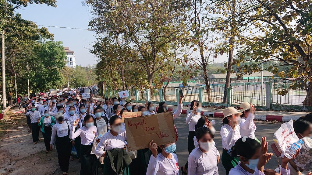 缅甸的教师在绿树成荫的道路上举着写着“尊重我们的选票”的标语牌游行。 行军向后延伸到远方。