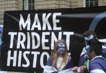 3 young people with saltires painted on their faces stand in front of a banner reading Make Trident History.
