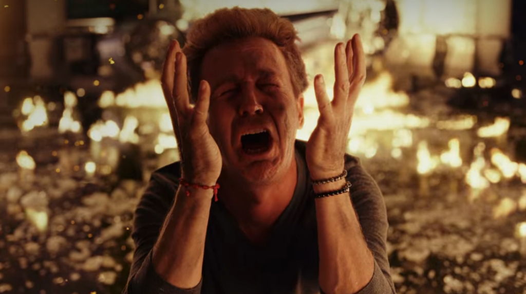 Изображение показывает Эдварда Нортона в роли его персонажа в «Стеклянном луке», в отчаянии, с поднятыми вверх руками и кричащим на фоне огня.