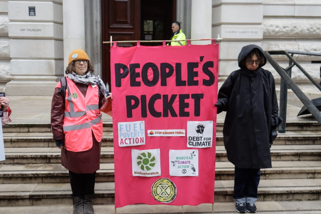 À l'extérieur du trésor, deux personnes tiennent une bannière rose sur laquelle on peut lire « Picket du peuple », avec le nom de différentes organisations cousu dessus.