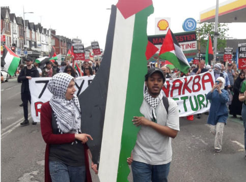 L'immagine mostra i manifestanti che camminano per Rusholme a Manchester con in mano una grande scultura della Palestina dipinta con i colori della bandiera palestinese.