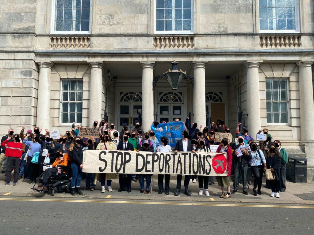 una grande folla si trova fuori dal tribunale con uno striscione con la scritta Stop Deportations