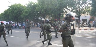 Police in green uniform and helmets and holding batons run down a road in Senegal. They are turning back to look at the protestors behind them.
