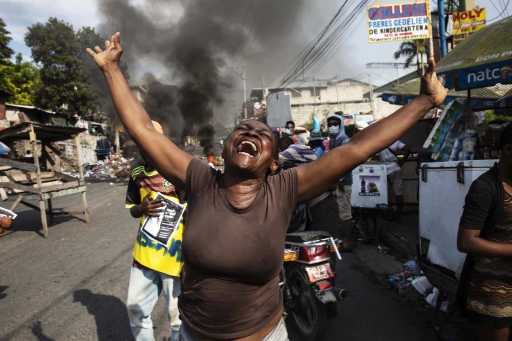 Une femme lève les bras en l’air lors d’une manifestation, tandis que de la fumée s’élève en arrière-plan. 